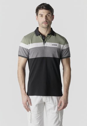 Hombre vistiendo una camiseta tipo polo de manga corta con rayas en verde oliva, blanco, gris y negro, y pantalones cargo blancos, de pie contra un fondo liso.