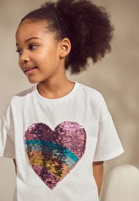 Fehér póló, amelyen egy nagy szív látható, amely több színű paillettesből áll, rózsaszín, kék és arany tónusokban. Rövid ujjú és lazán szabott.