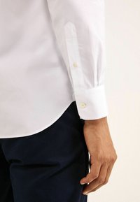 Weißes Langarmhemd mit spitzen Manschetten und zwei Knöpfen, kombiniert mit einer dunkelblauen Hose. Angenehmer Baumwollstoff, schlichtes Design.