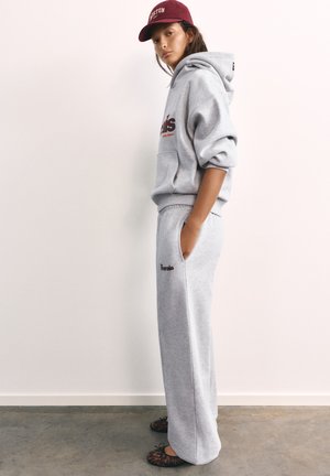 Jeune personne en sweat à capuche gris et pantalon de survêtement, les mains dans les poches, portant une casquette bordeaux et des ballerines à motifs, debout de profil.
