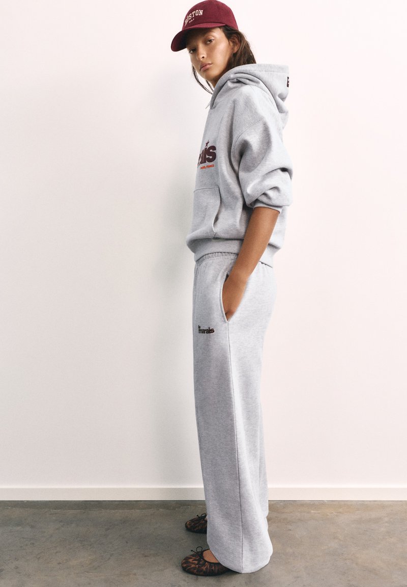 Jeune personne en sweat à capuche gris et pantalon de survêtement, les mains dans les poches, portant une casquette bordeaux et des ballerines à motifs, debout de profil.