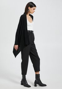 Cardigan negro abierto, camiseta ajustada blanca, pantalones negros de cintura alta, botas negras hasta el tobillo con un tacón grueso, detalles texturizados.