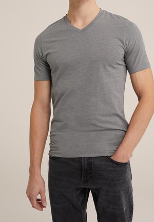 Man draagt een effen grijs V-hals T-shirt met korte mouwen en zwarte jeans, staat met één hand in de zak tegen een neutrale achtergrond.