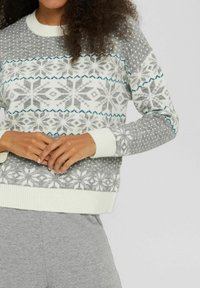 Person, die einen grau-weißen gestrickten Pullover mit Schneeflockenmustern und graue Hosen trägt, mit sanft gefalteten Händen in der Taille.