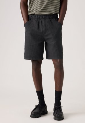 Musta tekstuuriga lühikesed püksid, millel on elastne vöökoht ja küljetaskud, kombineeritud mustade sokkide ja kingadega, kantud triibulise särgiga.