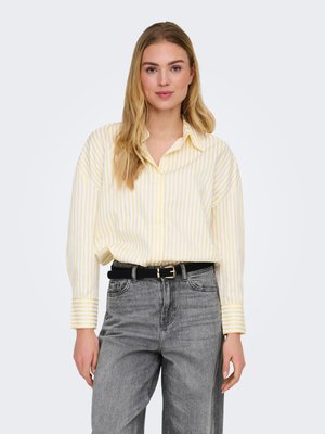 Vrouw met een geel-wit gestreept overhemd met knopen, ingestopt in een hoogwater grijze spijkerbroek met een zwarte riem, staand tegen een effen achtergrond.