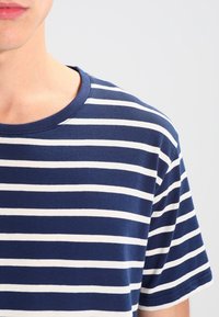 Bavlněné tričko s námořnicky modrými a bílými vodorovnými pruhy, s kulatým výstřihem a krátkými rukávy. Měkká textura a uvolněný střih.