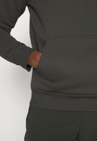 Felpa a maniche lunghe verde scuro con polsini a coste e tasca, mano parzialmente dentro la tasca, abbinata a pantaloni verde scuro coordinati.