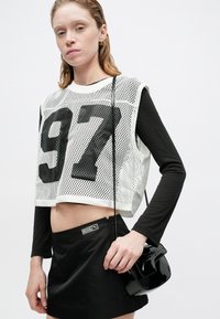 Witte mesh crop top met zwarte "97" print, gecombineerd met een zwarte mini rok en een kleine zwarte lakleren tas. Lange zwarte mouwen zichtbaar eronder.