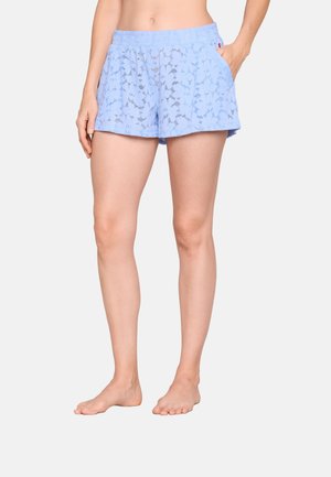 Femme portant un short à motifs bleu clair avec une taille élastique et des poches, debout pieds nus devant un fond uni.
