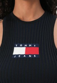 Black ribbed tank top featuring a rectangular logo with blue, white, and red accents, reading "TOMMY JEANS" in block letters.