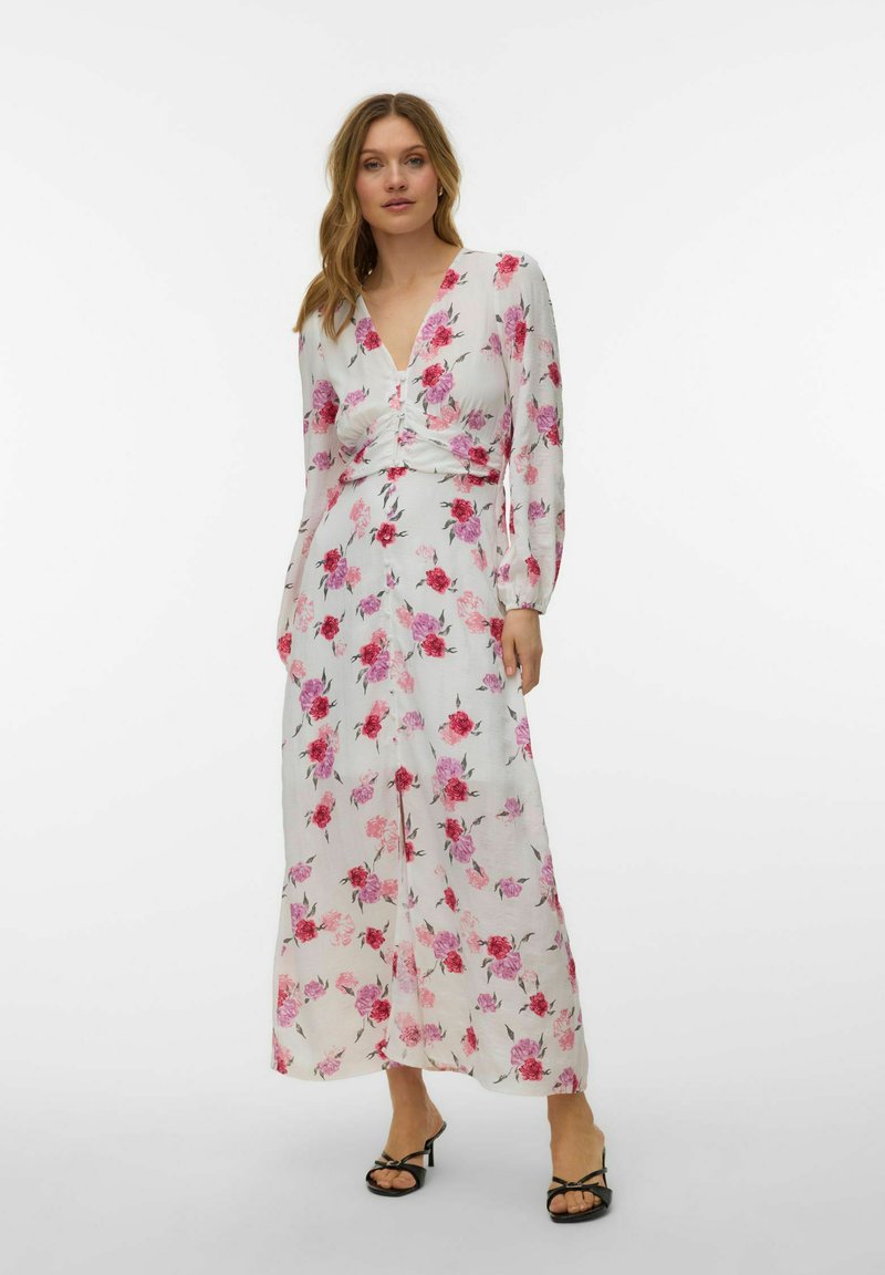 Femme portant une longue robe blanche à fleurs roses et rouges, manches longues, décolleté en V, et sandales noires à talons, debout sur fond blanc.