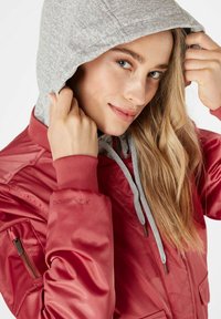 Chaqueta bomber roja con capucha gris. Cuenta con bolsillos con cremallera, puños acanalados y cordones. Textura suave con un acabado brillante.
