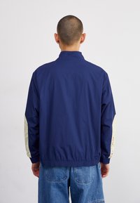 Marinblå vindjacka med krämfärgade ärmar, elastiska muddar och nederkant. Bakdelen har en enkel design och tygstruktur.