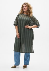 Olivgrünes Mesh-Kleid mit kurzen Ärmeln und lockerer Passform, mit gestreiften Akzenten am Halsausschnitt und an den Bündchen. Kombiniert mit blauen Jeans.