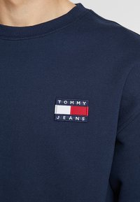 Marineblauwe sweatshirt van katoen, met een gestikt rechthoekig logo-embleem met "TOMMY JEANS" in wit en rood op de linkerborst.