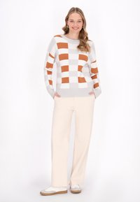 Creme- und orangefarbener karierten Pullover mit Rundhalsausschnitt, geripptem Saum und langen Ärmeln, getragen mit hellbeigen weit geschnittenen Hosen und weißen Sneakers.