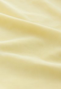 Pehme vaalea keltainen kangas, jossa hieno sileä rakenne ja lempeät laskokset, viitaten kevyeseen ja hengittävään materiaaliin.
