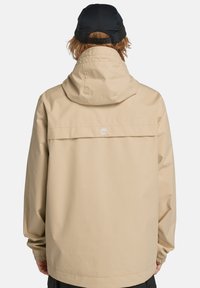 Beige vattentät jacka med huva, med ryggsömn och elastiska ärmslut. Logotyp broderad på övre delen av ryggen. Enkel, avslappnad passform.