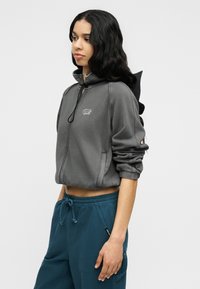 Γυναίκα με μακριά μαύρα μαλλιά, που φοράει γκρι ζακέτα με φερμουάρ και μπλε παντελόνι με κορδόνι, στέκεται προφίλ μπροστά σε απλό φόντο.