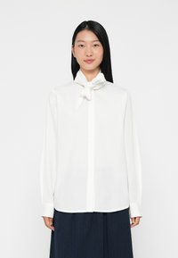 Camicia bianca a maniche lunghe con colletto a farfalla annodato, taglio sartoriale e texture liscia. Chiusura con bottoni frontale e ai polsini, abbinata a una gonna blu navy.