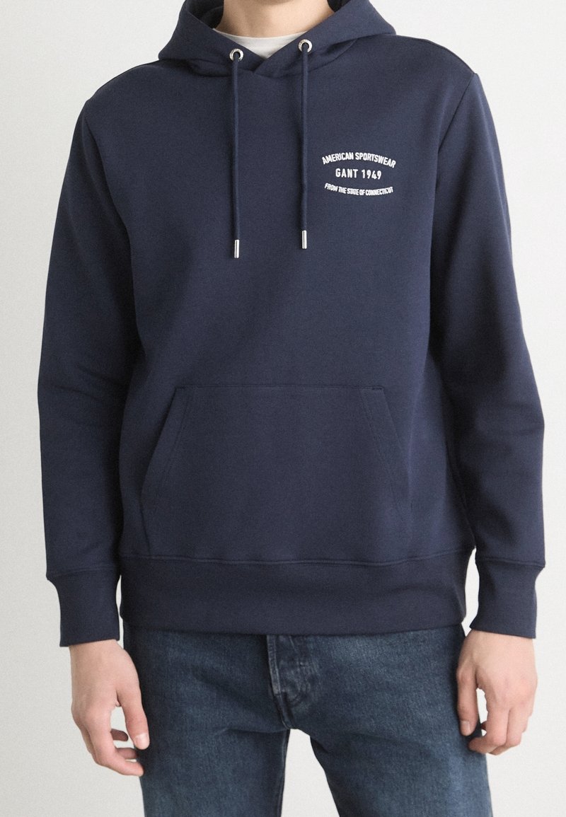 Homme portant un sweat à capuche bleu marine avec poche avant et texte blanc « American Sportswear GANT 1949 From the State of Connecticut » sur la poitrine.