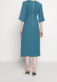 Robe midi bleu-vert avec des manches mi-longues, une taille cintrée et une fermeture éclair au dos. Tissu au fini lisse et jupe légèrement évasée.