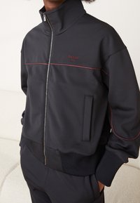 Giacca nera con zip in tessuto elasticizzato, caratterizzata da cuciture rosse e colletto alto. Include una tasca laterale e polsini a costine.