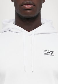 Sweatshirt branco com capuz, apresentando um tecido texturizado, cordões ajustáveis e um pequeno logótipo bordado a preto no lado esquerdo do peito.