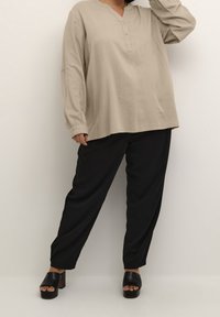 Beżowa koszula z długim rękawem, dekoltem w serek i detalem guzików, w połączeniu z czarnymi szerokimi spodniami. Modelka nosi czarne sandały na masywnym obcasie.