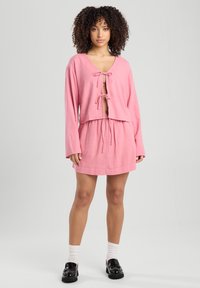 Camicia corta rosa chiaro con maniche lunghe e chiusura a fiocco, abbinata a una gonna con coulisse coordinata. Texture in cotone liscia.