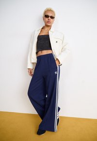 Navy blue wide-leg pants with white side stripes and adidas logo. Paired with a black cropped top and white jacket, worn with black shoes.