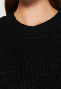 Gros plan sur le cou et le haut de la poitrine d'une personne portant un pull en maille côtelée noire avec un col rond.