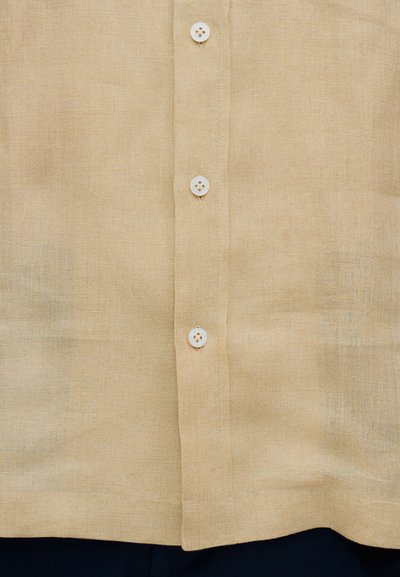 Chemise en lin beige avec une texture lisse, dotée d'un plastron à boutons et de boutons en plastique blanc. Design simple et ajusté.
