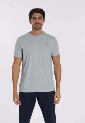 T-shirt gris à manches courtes en tissu doux, avec un col rond et un petit logo sur la poitrine. Le modèle l'associe à un pantalon marine.