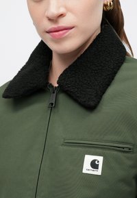 Groene jacket met een zwarte, gestructureerde fleece kraag, ritsluiting en een borstzak met een witte merkembleem.