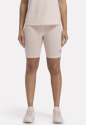 Person iført lyserøde Reebok cykelshorts, en matchende lyserød top og lyserøde sneakers, stående med hænderne afslappet langs siden.