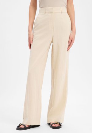 Mujer vistiendo pantalones anchos de cintura alta beige y sandalias planas negras, de pie frente a un fondo blanco liso.