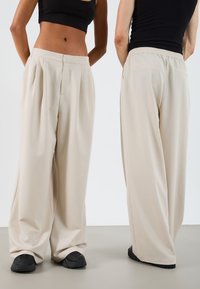 Pantalons plissés beige en tissu lisse ; dotés d'une taille élastique et de poches latérales ; design à jambes larges avec une coupe décontractée.