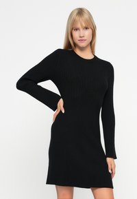 Donna bionda che indossa un abito a maniche lunghe in maglia a coste nera aderente, con una mano sulla vita, su uno sfondo bianco semplice.