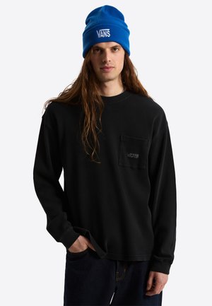 Junger Mann mit langen Haaren, der eine blaue Vans-Mütze und ein schwarzes Langarmshirt mit Brusttasche und Vans-Logo trägt.