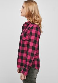 Chemise à carreaux rose et noir avec un col pointu, une poche poitrine unique et des manches longues. Tissu texturé, détail d'ourlet fendu sur les côtés.