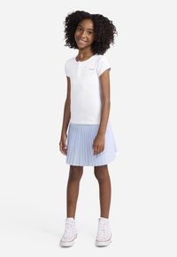 Λευκό κοντομάνικο πουκάμισο με γιακά henley, συνδυασμένο με φούστα με πτυχώσεις σε ανοιχτό μπλε χρώμα. Το σύνολο ολοκληρώνουν λευκά παπούτσια τύπου high-top.