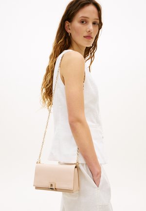 Young woman with long wavy hair wearing a white sleeveless top and pants, carrying a beige crossbody bag with a gold chain strap.