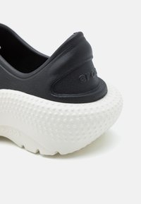 Svart slip-on sko med en glatt, teksturert overdel og en kraftig hvit såle med et prikket mønster. Logo synlig på hælen.