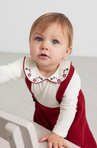 Rotes Corduroykleid mit einem weißen Langarmhemd, das einen gewellten Kragen hat und mit floraler Stickerei verziert ist. Kind, das nach einem Tisch greift.