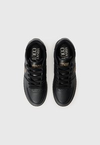 Baskets en cuir noir avec des panneaux texturés, des lacets plats noirs, et une marque "Versace Jeans Couture" dorée sur le côté.