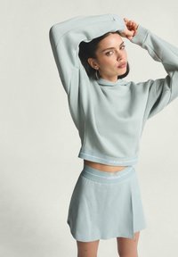 Svetlomodrý skrátený hoodies a zodpovedajúca sukňa vyrobená z jemnej látky, s vyšitým logom pozdĺž lemu a pásu.