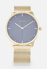 Reloj con correa de malla dorada y esfera redonda en azul-gris. Presenta marcaciones minimalistas, agujas doradas y un logo de Calvin Klein. Diseño simple y moderno.