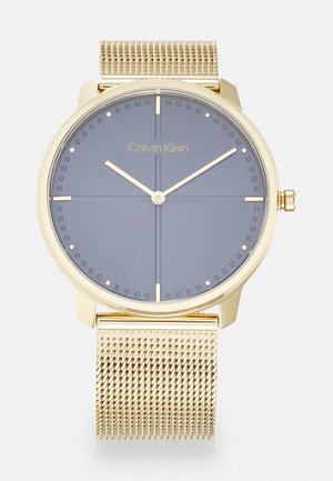Montre avec bracelet en maille dorée et cadran rond bleu-gris. Elle présente des marquages minimalistes, des aiguilles dorées et un logo Calvin Klein. Design simple et moderne.